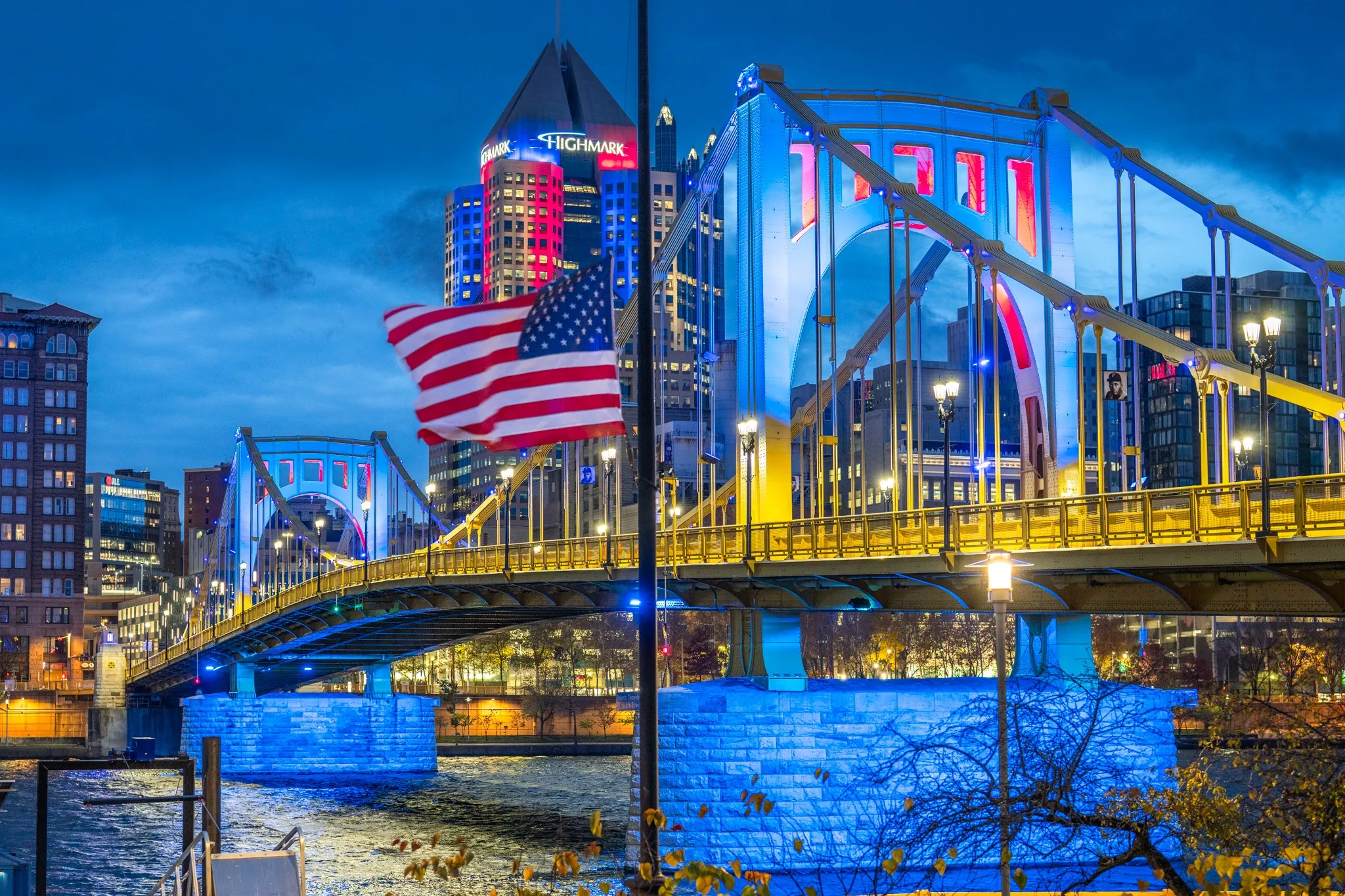 An American Flag flies on a Pittsburgh bridge at dusk, the lit downtown skyline behind it.
